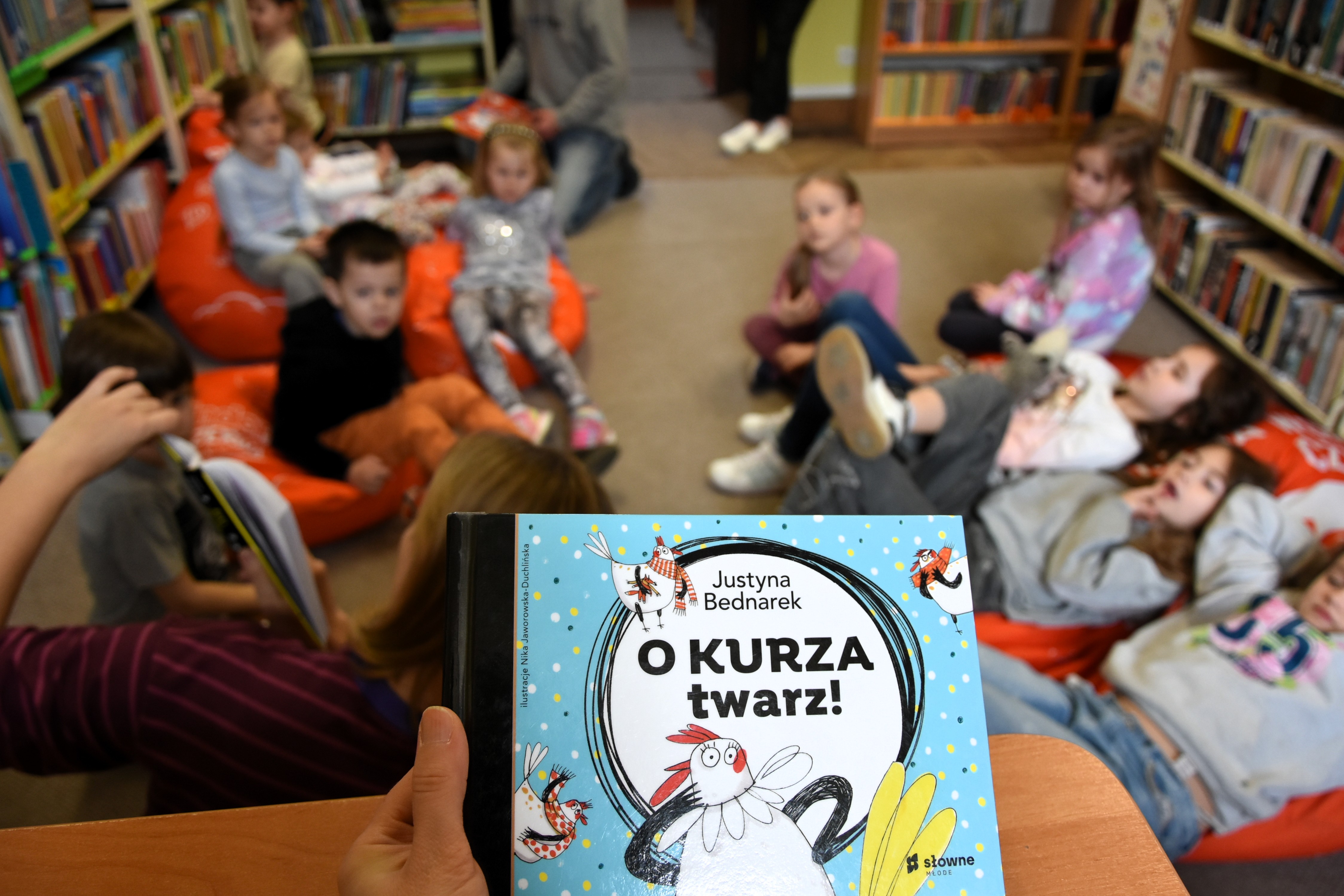 Na pierwszym planie książka ,,Kury z grubej rury", w tle dzieci siedzące na pufach