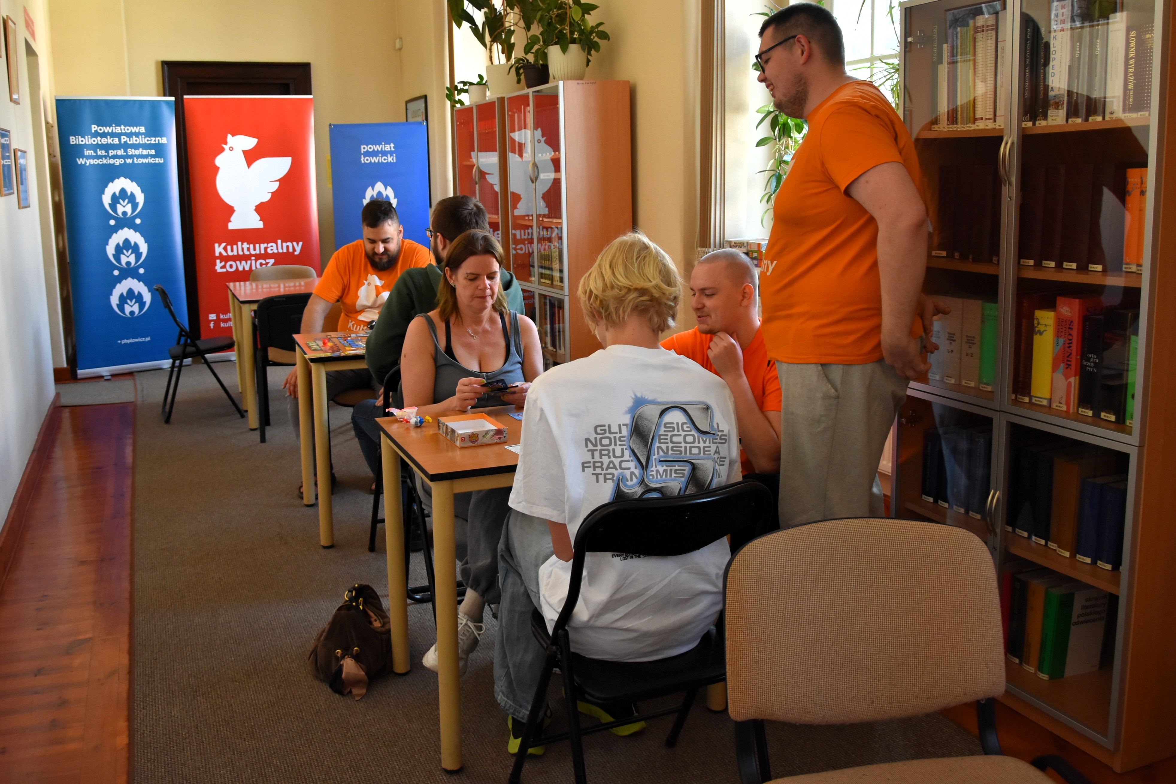 Grupa osób siedzi przy stolikach i gra w planszówki. W tle regały z książkami i banery.